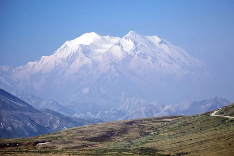 Snow-capped mountain peaks rise above rolling hills with patches of grass and a winding road in the foreground, under a clear blue sky.