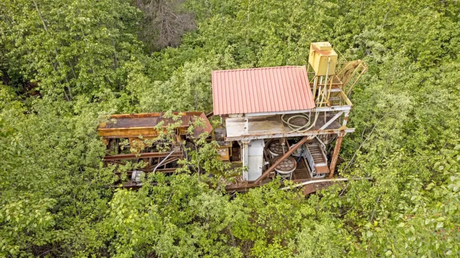 A rusted, abandoned mining machine with a red metal roof is surrounded and partially overgrown by dense green vegetation. The machinery appears weathered and entangled in the thick foliage.
