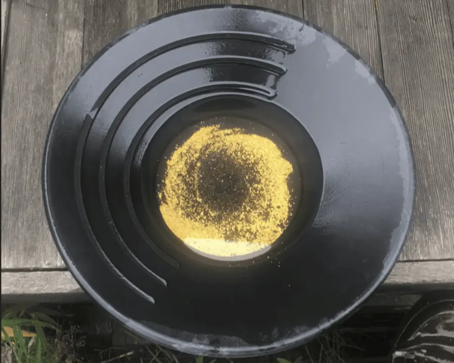 A black gold pan containing a mix of gold flakes and black sand, resting on a wooden surface outdoors.