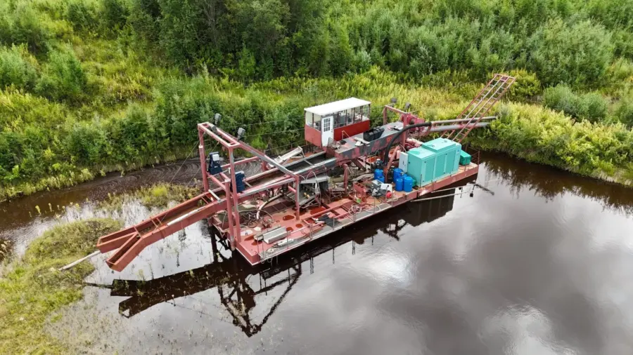 A red and white floating dredge with equipment and storage containers sits on a calm, narrow body of water, surrounded by dense green vegetation and trees.