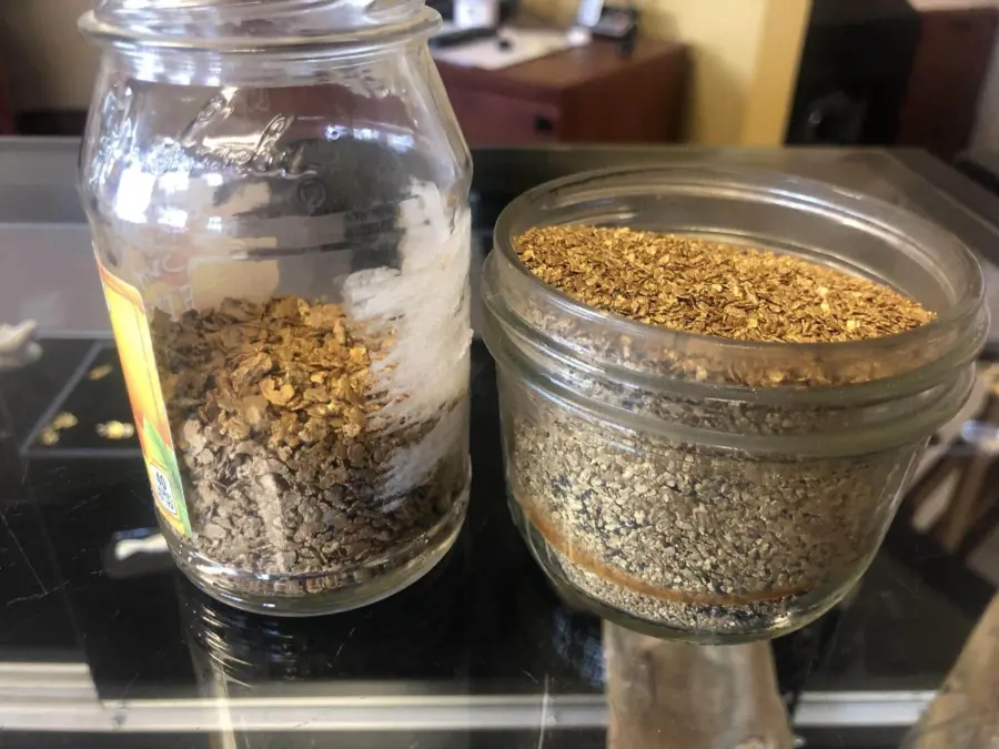 Two small glass jars filled with brown, flaky dried herbs or plant material sit on a reflective surface. One jar is tall with a label and the other is shorter and wider.