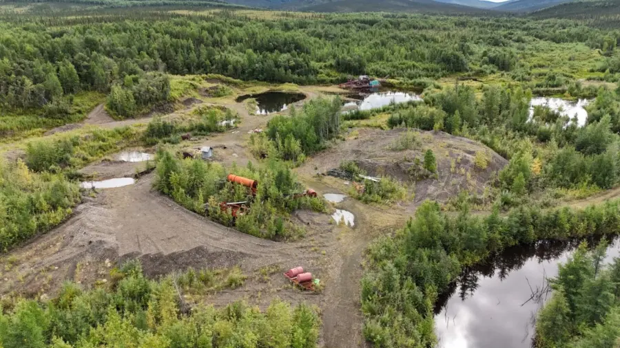Aerial view of an old, overgrown mining site with scattered rusted equipment, dirt piles, small ponds, and dense green forest surrounding the area under a cloudy sky.