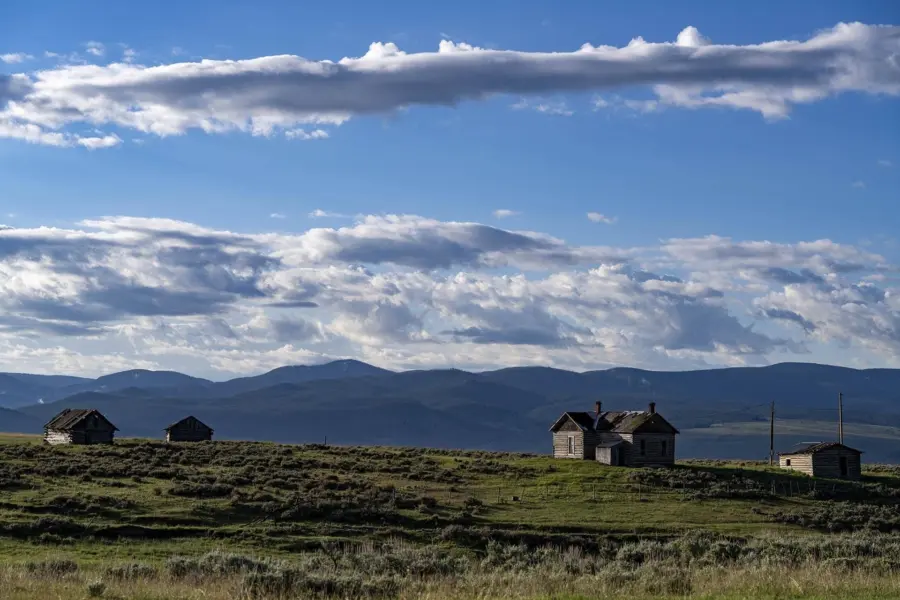 Several small wooden cabins sit on a grassy plain under a blue sky with scattered clouds, with distant mountains visible along the horizon.