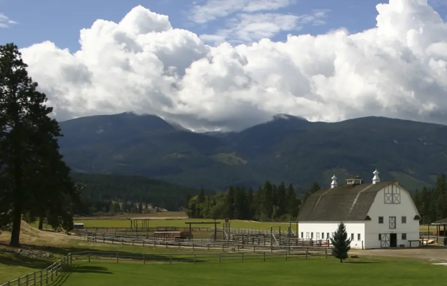 A white barn with a dark roof stands in a green field, surrounded by wooden fences, with forested hills and tall mountains in the background under a sky filled with large, fluffy clouds.