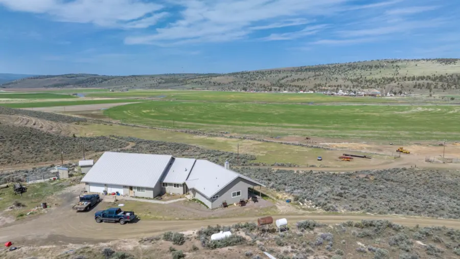 Aerial view of a beige house with a metal roof and several vehicles parked outside, situated in a rural landscape with green fields, hills, and scattered buildings under a blue sky with clouds.