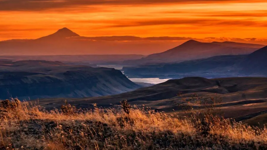 Rolling hills with dry grass in the foreground, a river winding through canyons, and distant mountains under a vivid orange sunset sky.