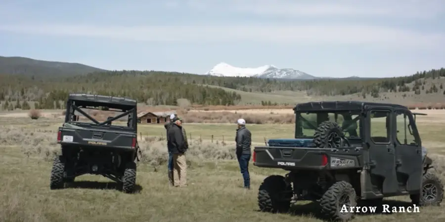 Two people stand in a grassy field beside two Polaris off-road vehicles, looking toward distant snow-capped mountains and a cabin. The words Arrow Ranch appear in the lower right corner.