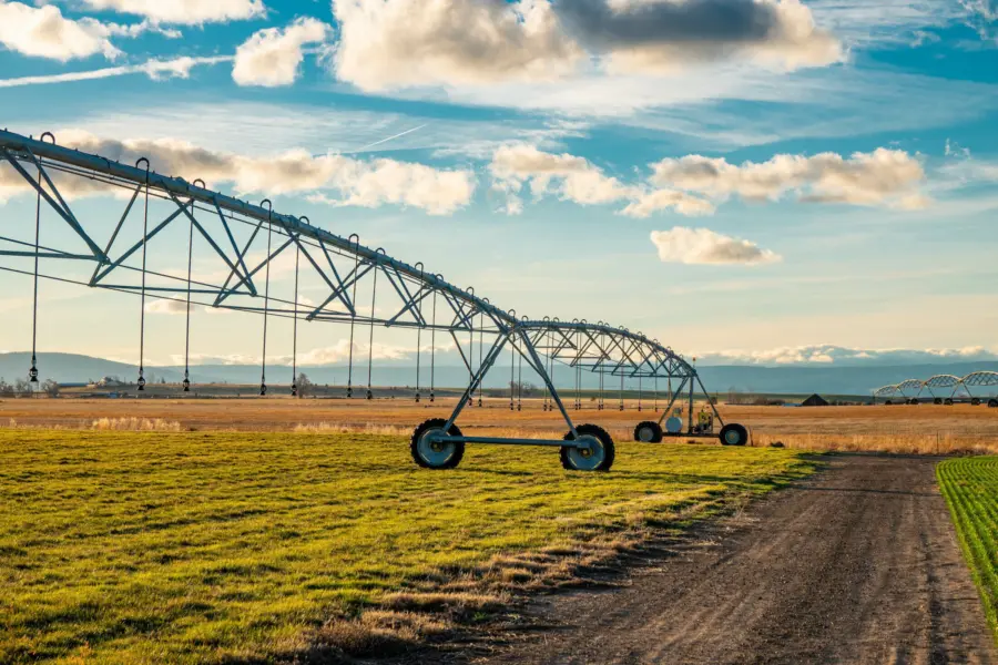 A large irrigation system with wheels stretches across a green and brown field under a partly cloudy sky, with mountains visible in the distance and a dirt path running alongside the equipment.
