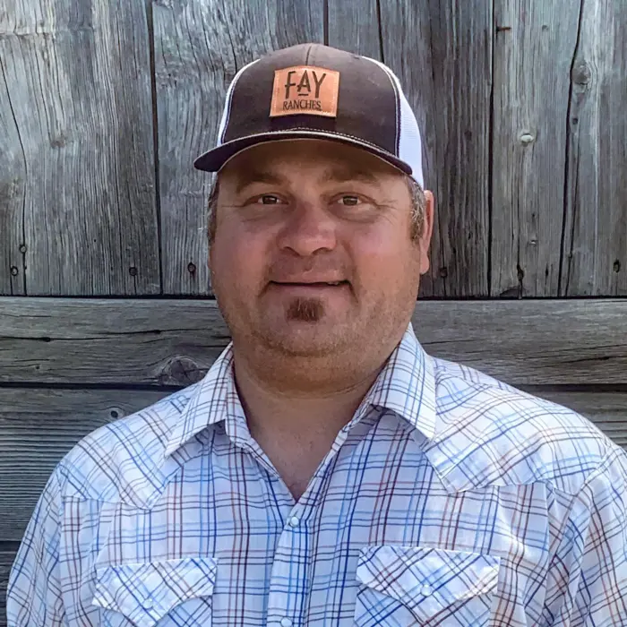 A man wearing a plaid shirt and a black-and-white baseball cap that says Fay Ranches stands in front of a weathered wooden wall, smiling slightly at the camera.