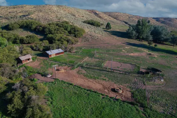 Aerial view of a rural farm with three wooden barns, fenced animal pens, and patches of green grass surrounded by hills and scattered trees under a partly cloudy sky.
